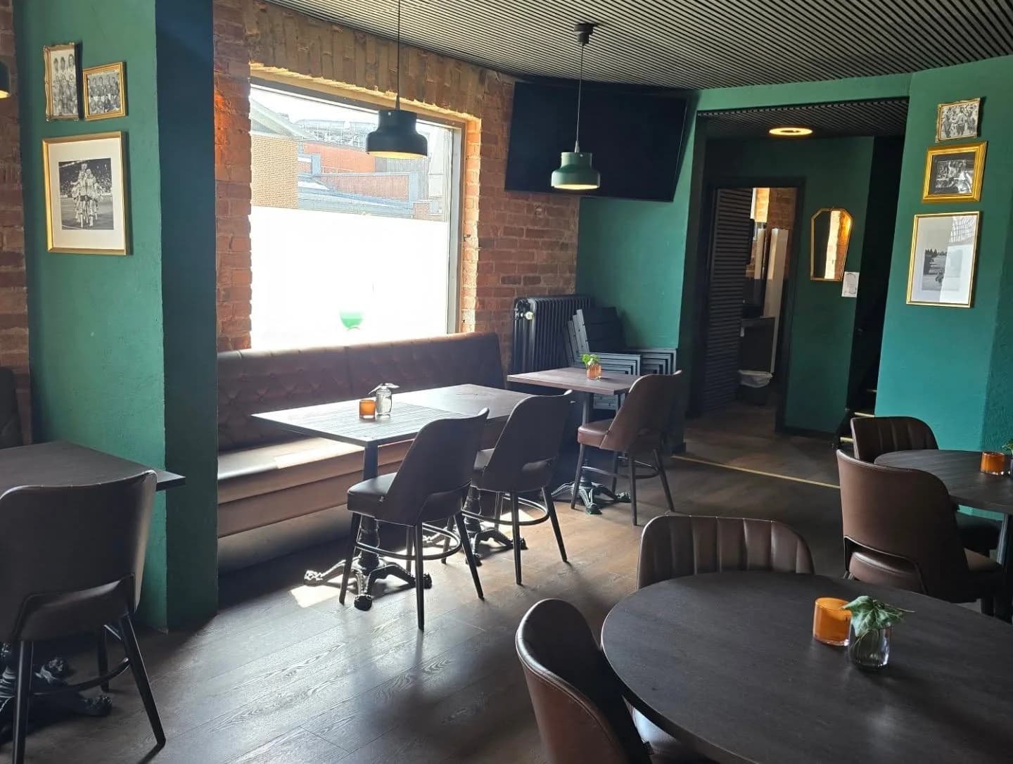 Läderbänk under ett fönster i tegelväggen, mörka bord med bruna stolar och inramade fotbollsfoton på gröna väggar